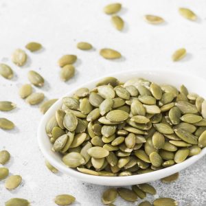 Image of roasted pumpkin seeds in a white bowl with pumpkin seeds scattered all around the bowl as well.