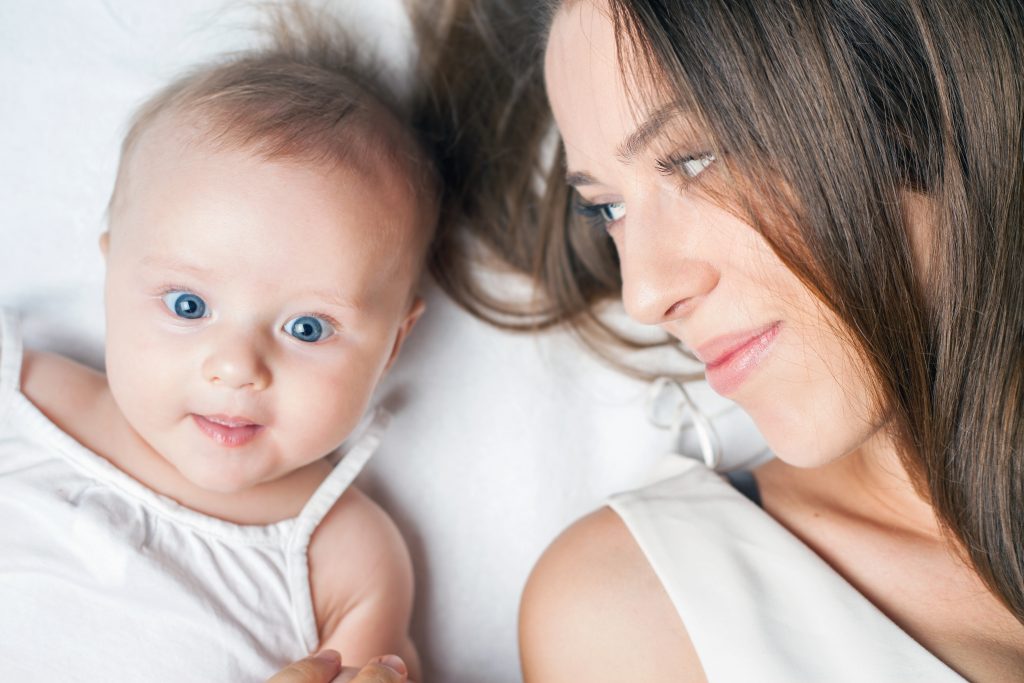 Happy baby girl lying near her mother on a white bed. Newborn looking at the camera and smiling. Mothercare is most important in baby life