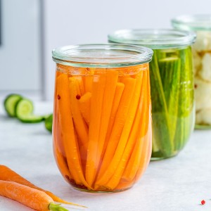 Three clear glass jars; one with pickled carrots, one with pickled cucumbers, and one with pickled cauliflower.