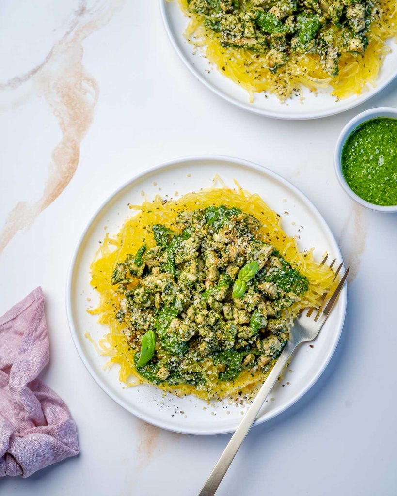 Vibrant green pesto sauce on chicken topping a mound of orange spaghetti squash next to a fork on a white plate.