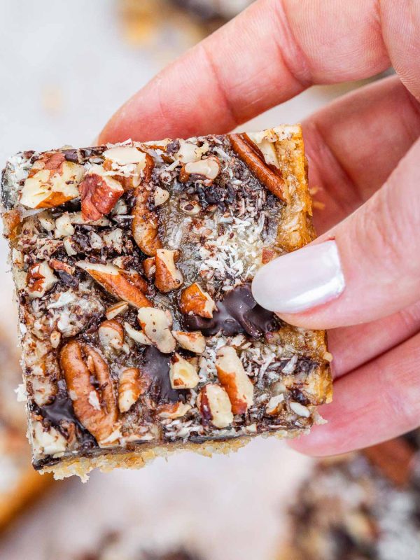 A hand with white fingernails holding a cookie bar topped with chocolates and nuts.