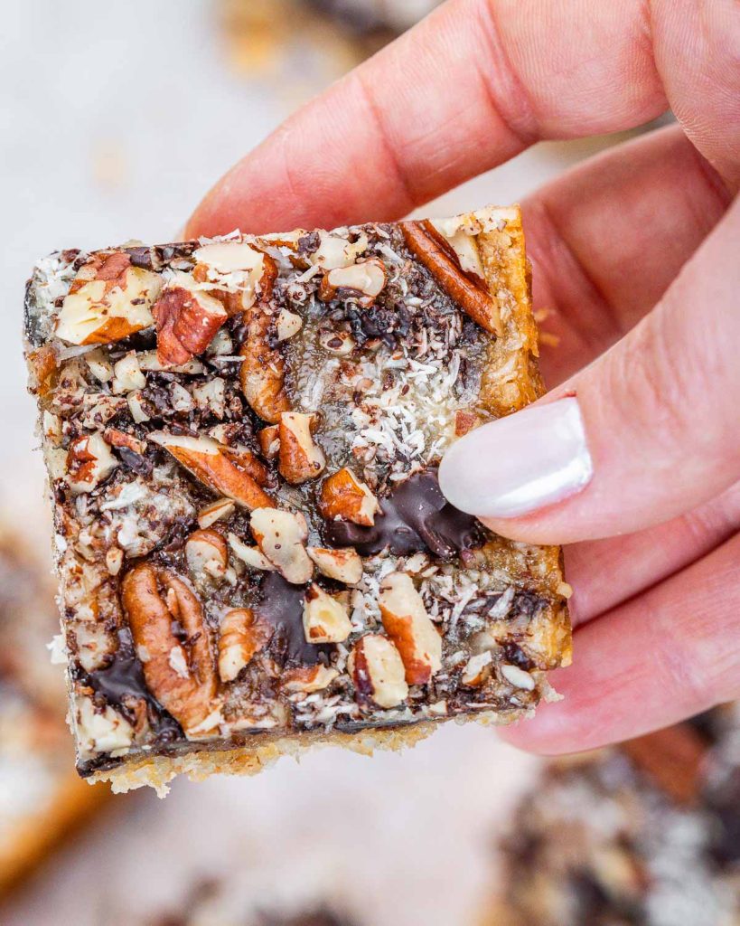 A hand with white fingernails holding a cookie bar topped with chocolates and nuts.