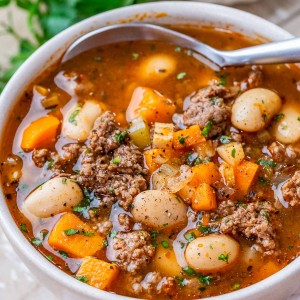 Beef vegetable soup with beans, carrots, and ground beef and a silver spoon in a white bowl.