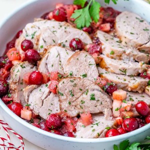 Sliced pork loin in a white bowl covered with cranberries and apples.