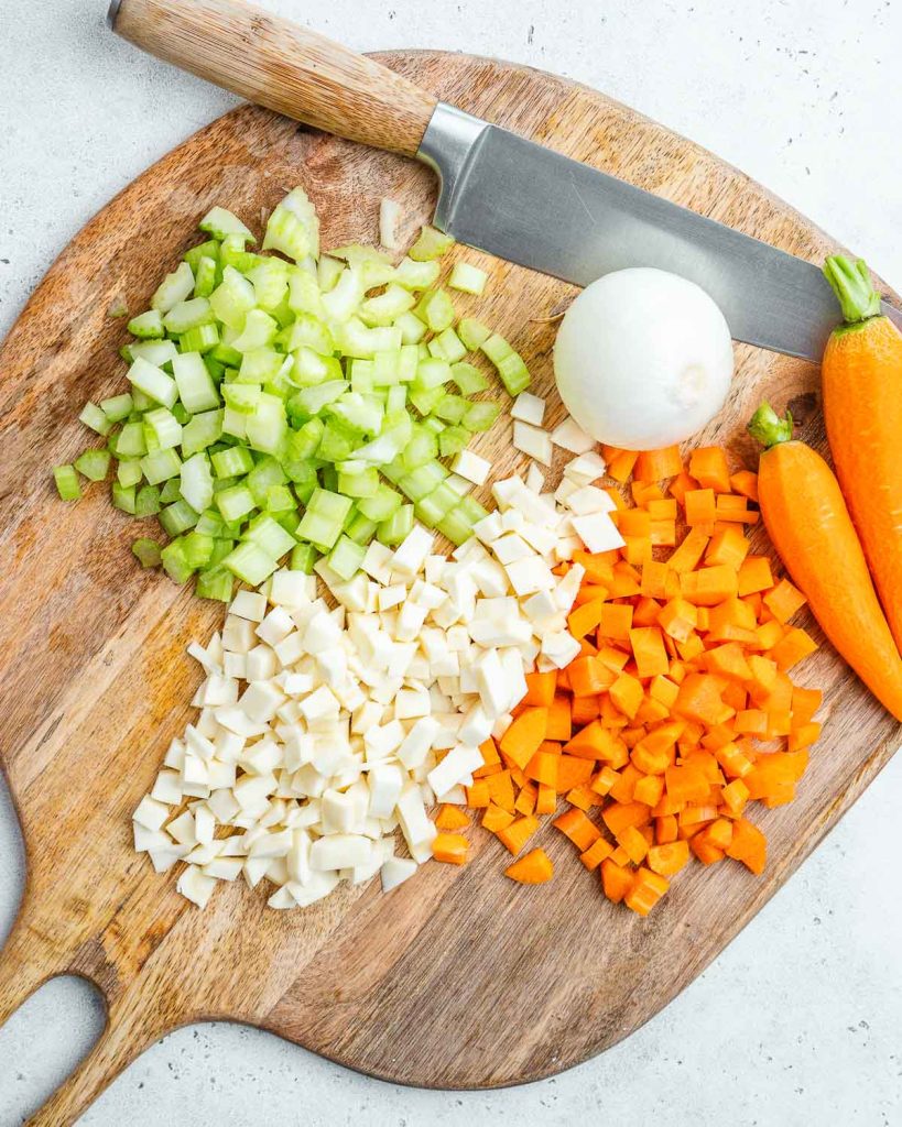 Chopped celery, parsnip, and carrots on at cutting board next to a knife.