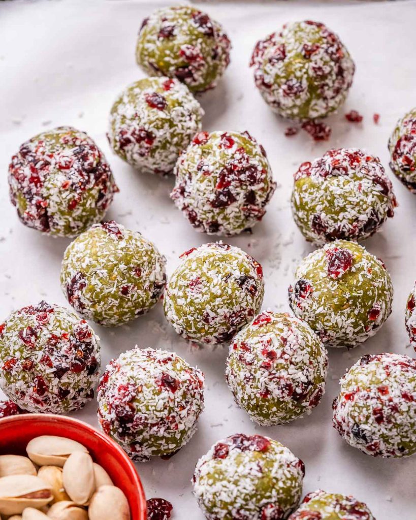 Green, red and white colored Christmas Energy Balls laid out on a white background next to a red bowl of pistachios.