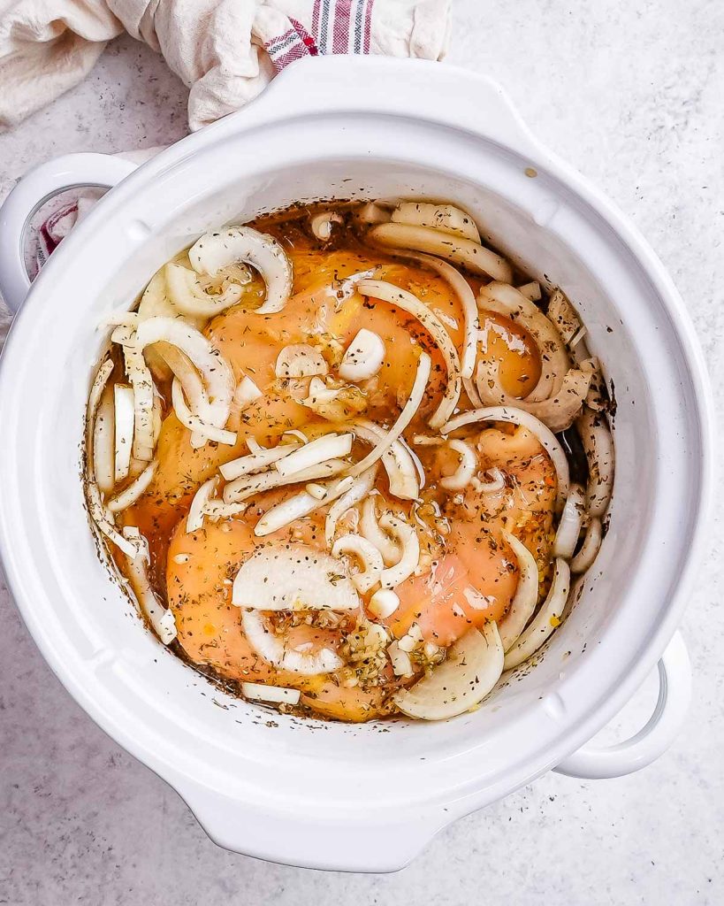 A white crock pot filled with chicken breast and sliced onions.