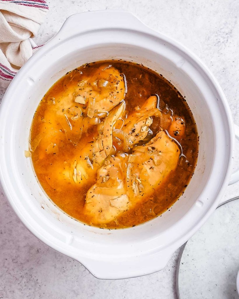 A white slow cooker containing broth and chicken breasts.