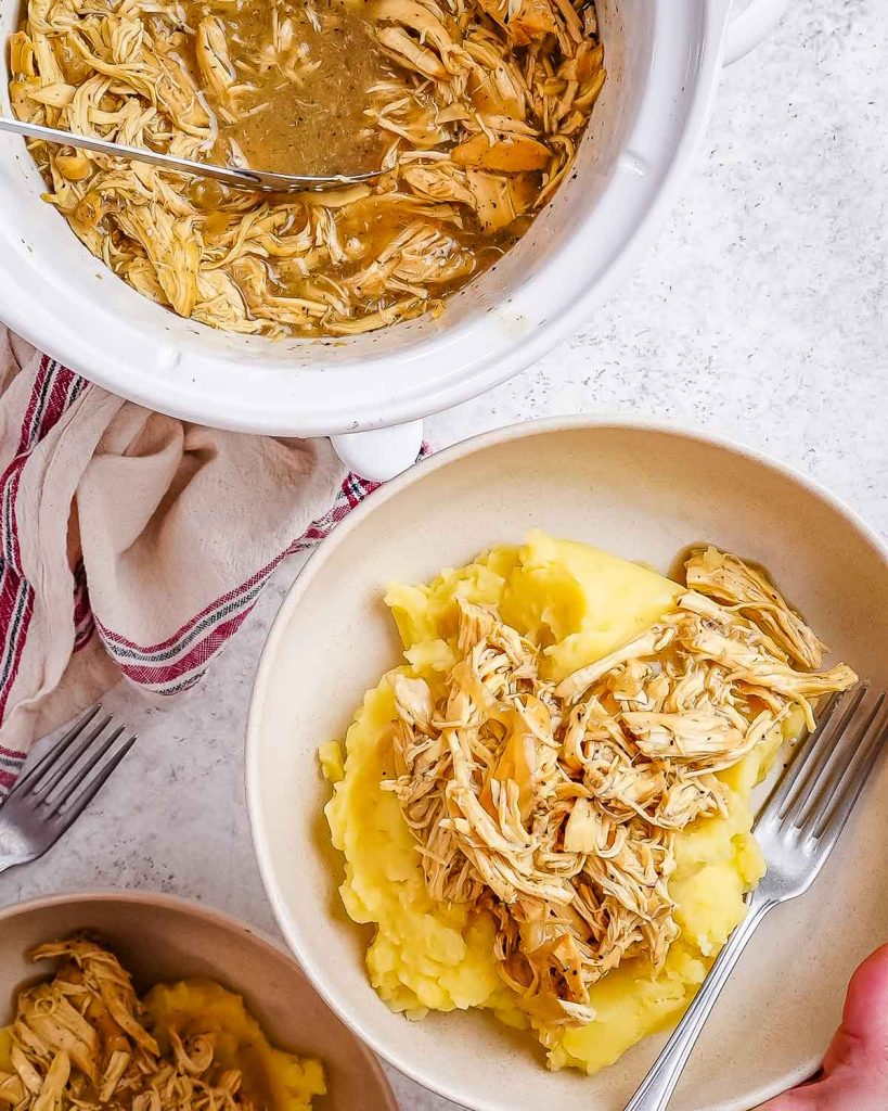 White slow cooker filled with shredded chicken next to a plate of mashed potatoes topped with shredded chicken.