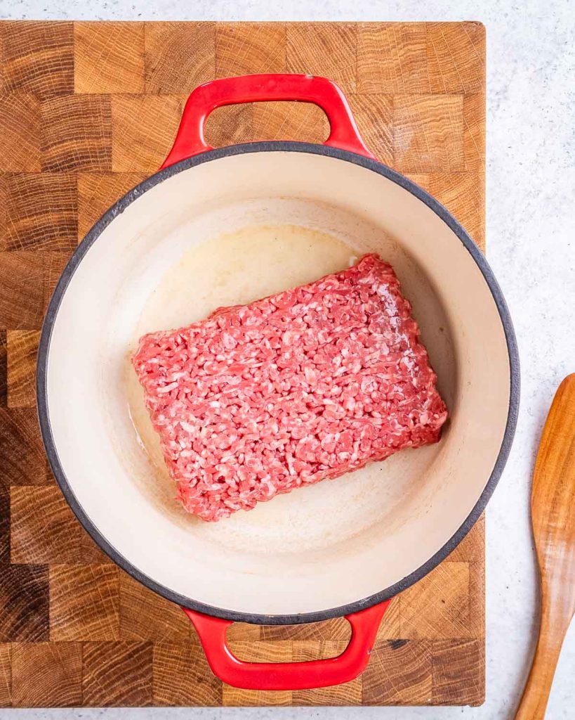 A rectangle of ground beef in a white enameled cast iron stock pot with red handles on a butcher block.