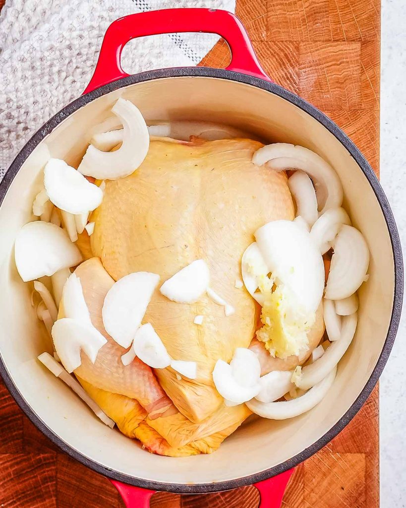 A whole raw chicken covered with onion slices and garlic in an enameled Dutch oven with red handles