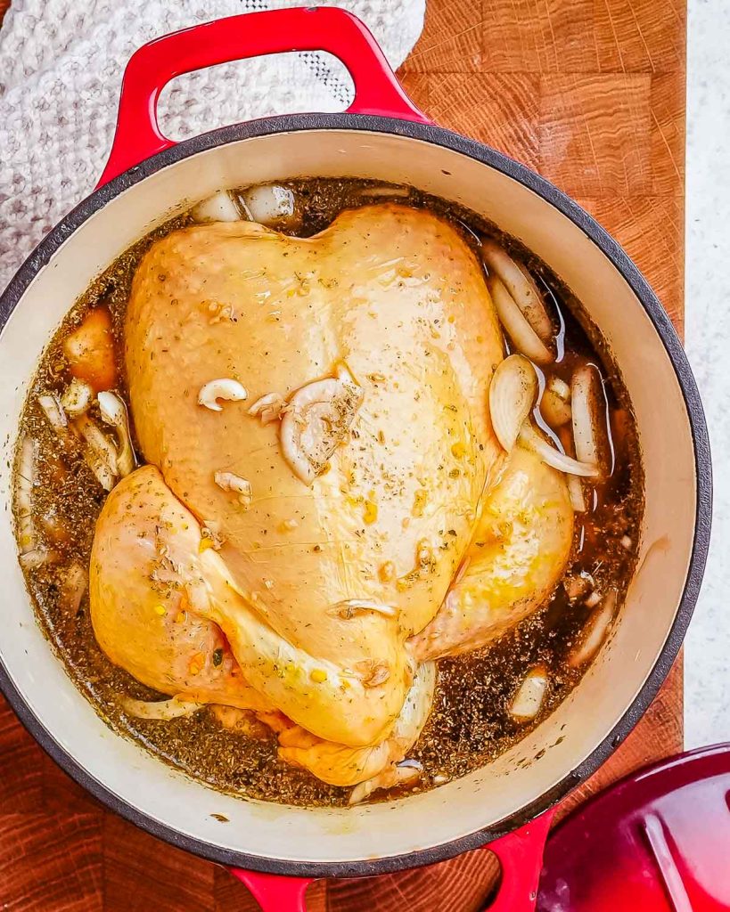 A whole chicken in a cast iron Dutch oven along with balsamic vinegar, seasonings, bone broth, and rosemary sprigs.