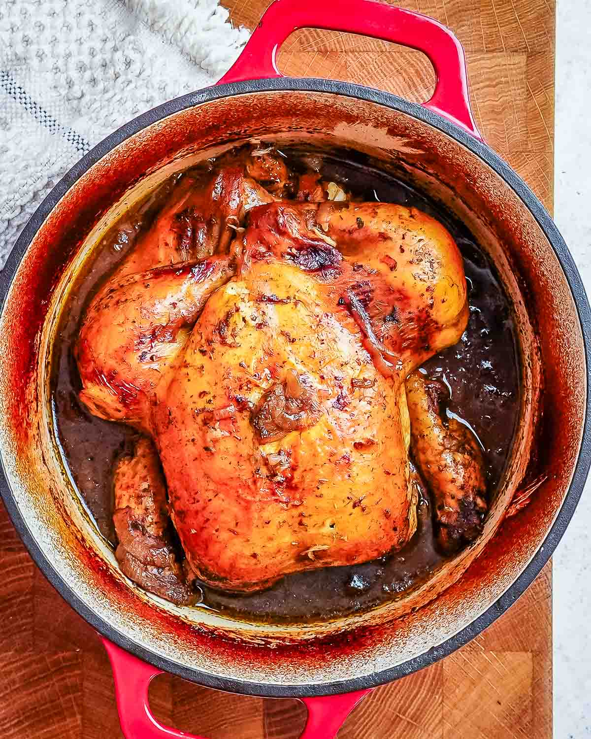 A large deep Dutch oven with red handles containing a whole baked chicken in a dark balsamic gravy.
