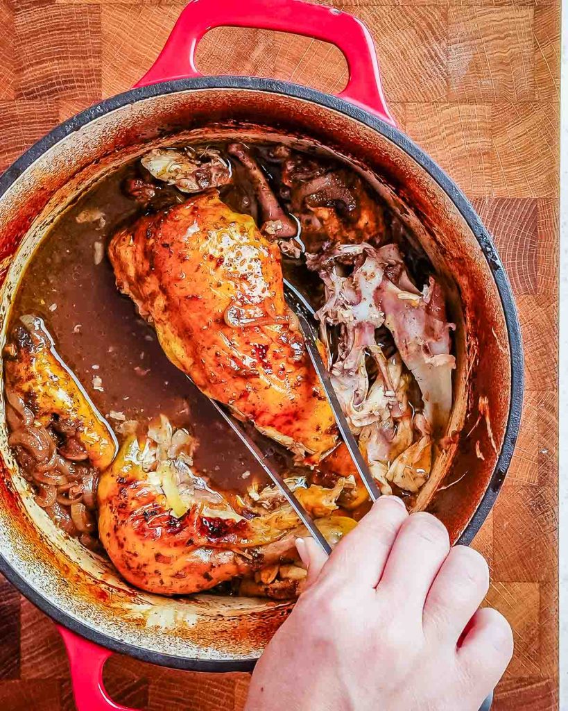 A cooked whole chicken being removed from a Dutch oven by a hand holding a pair of silver tongs.