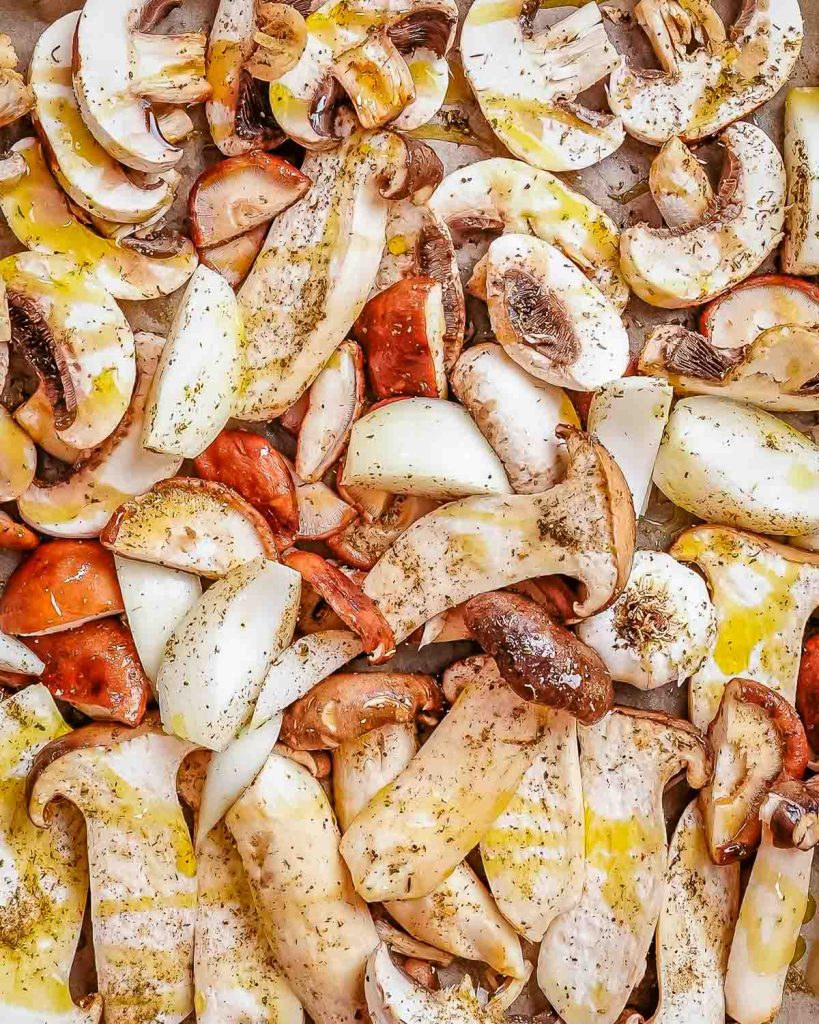 an assortment of chopped mushrooms, onions, and a bulb of garlic on a large sheet pan drizzled with olive oil.