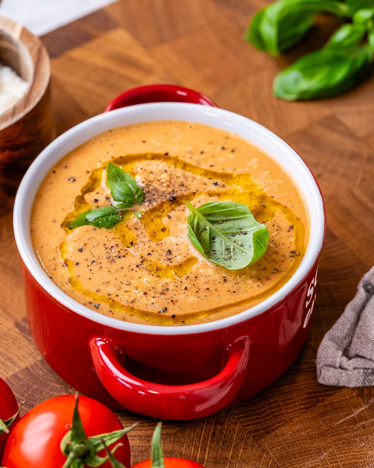 High protein roasted tomato soup garnished with basil leaves and a drizzle of olive oil in a red bowl with red handles.