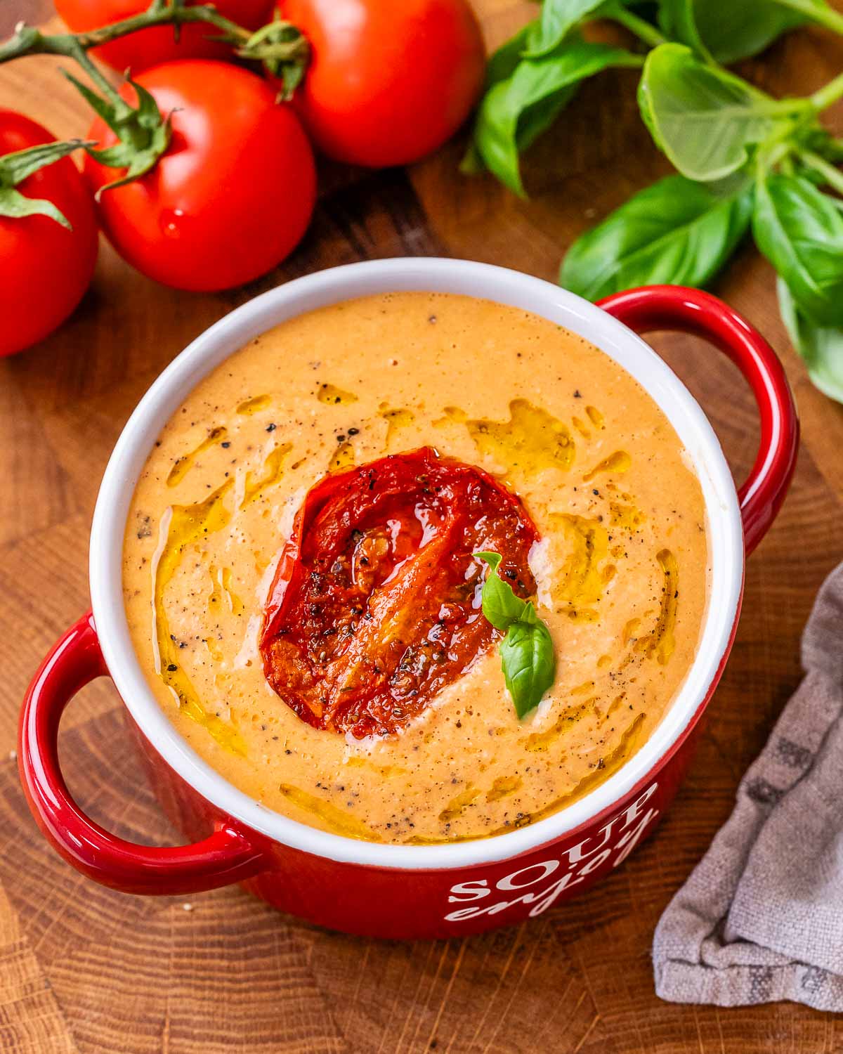 A bowl of roasted tomato soup containing chicken and cottage cheese garnished with a roasted tomato and small basil leaf and sitting on a brown cutting board next to red tomatoes and fresh green basil.