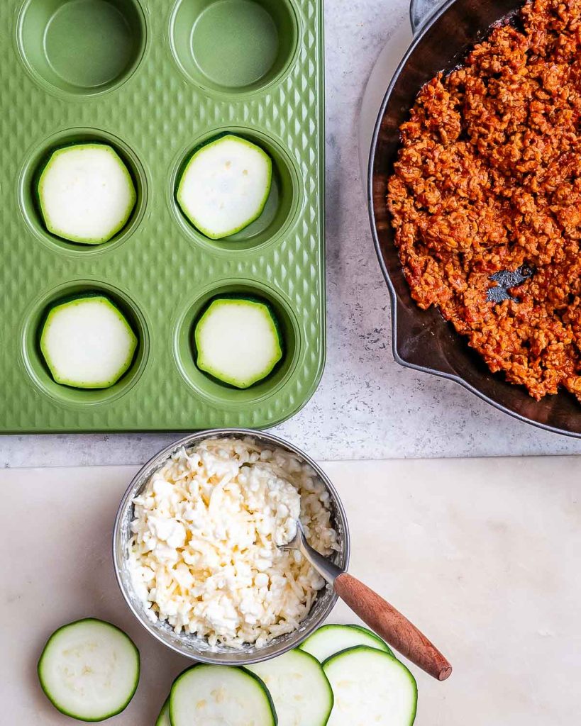A green silicon muffin tin with slices of zucchini in the bottom of the holes and a bowl of cheese mixture next to cooked beef in marinara.