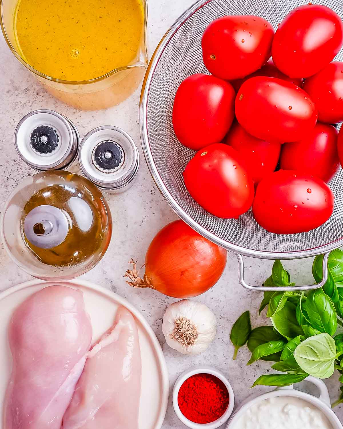 All the ingredients needed to make high protein roasted tomato soup laid out on a white and grey background: tomatoes, bone broth, salt, pepper, olive oil, onion, garlic, paprika, cottage cheese, chicken.