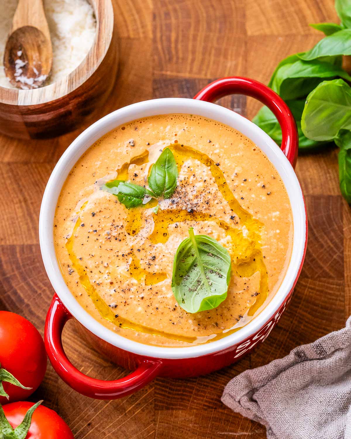 Creamy, orange colored high protein tomato soup in a bowl with red handles and topped with basil leaves and a drizzle of olive oil.